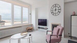 A modern living room with a large window, a flat-screen TV, a fireplace, and a clock on the wall. - Family Apartment in Ostend Near the sea (Ostend)
