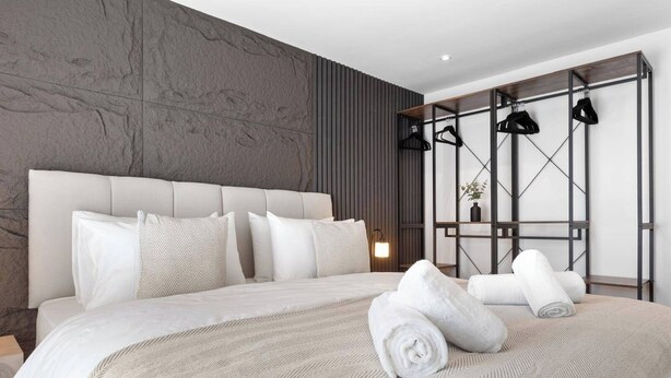 A modern hotel room with a large bed, a wooden panel wall, and a metal rack with hanging clothes. - Unique 2BR Finchley Flat Sleeps 5 (London)