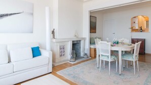 A modern living room with a white sofa, a fireplace, a dining table with chairs, and a patterned rug. - Studio With sea View in Ostend (Ostend)