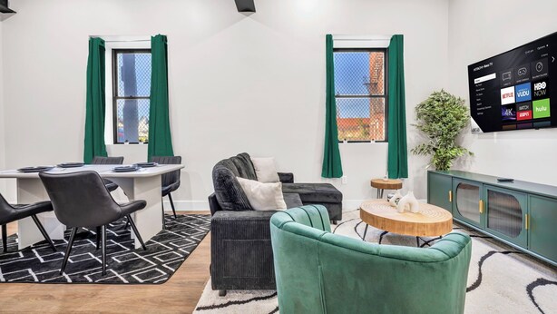 Ein modernes Wohnzimmer mit Essbereich, einem an der Wand befestigten Fernseher und Blick auf die Stadt durch die Fenster.