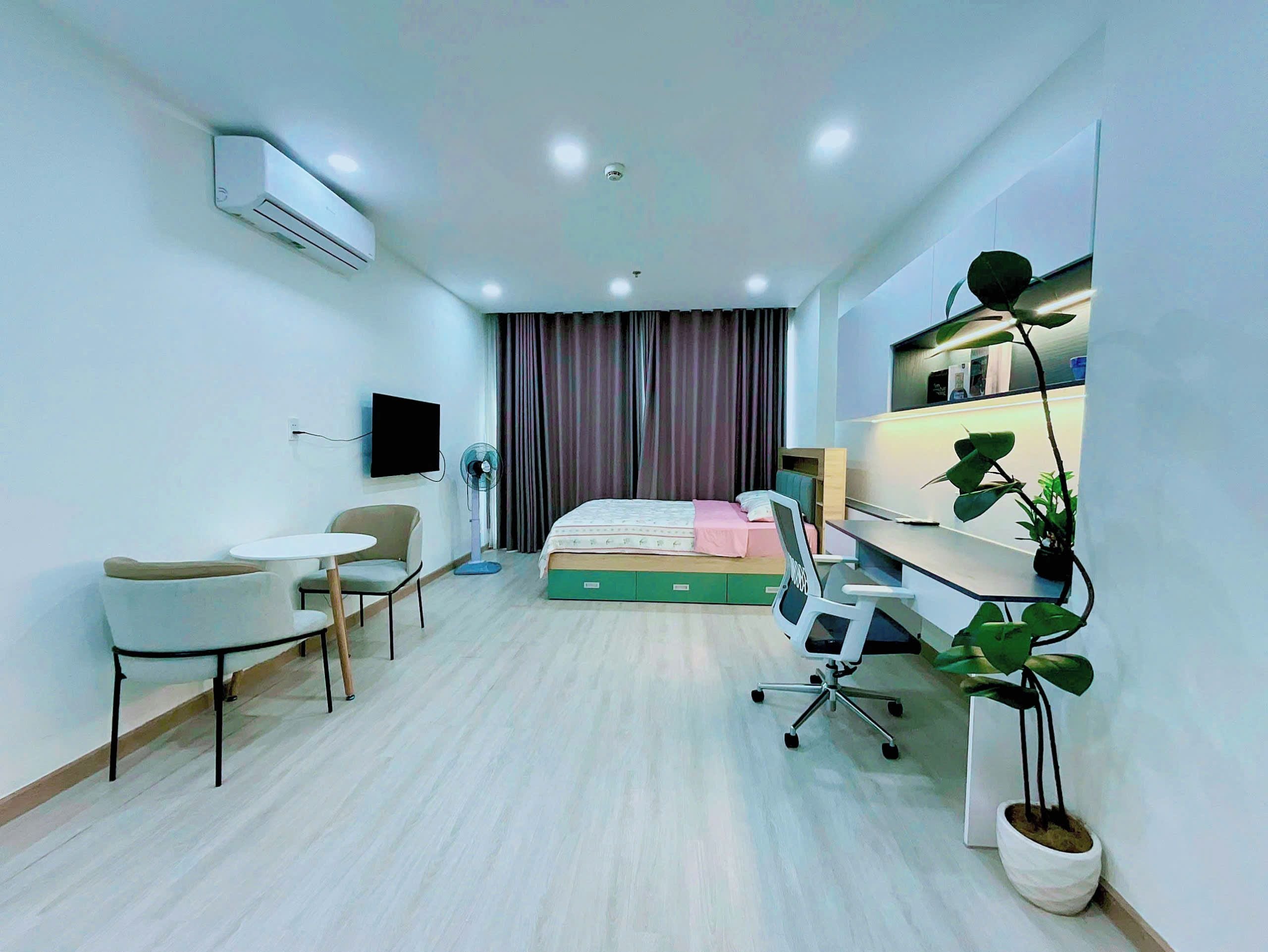A modern hotel room with a bed, desk, chair, air conditioning unit, TV, and potted plants.