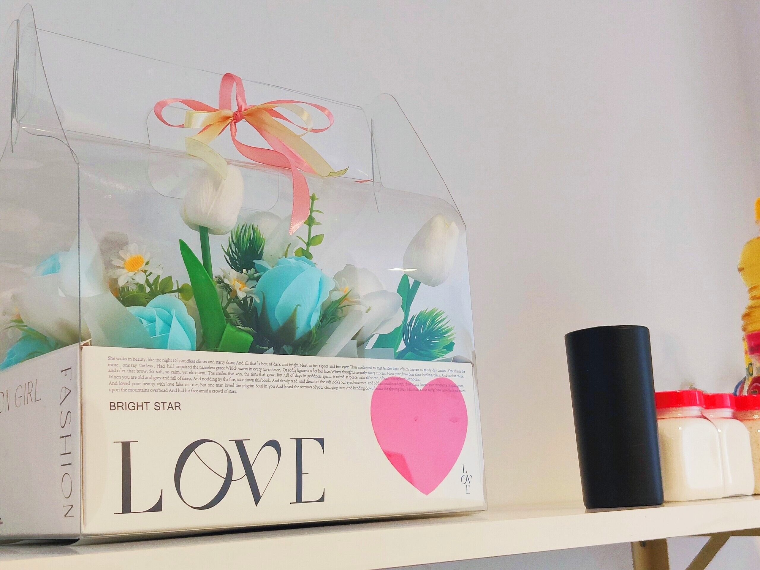 A clear plastic box with a pink ribbon, labeled "BRIGHT STAR LOVE," containing artificial flowers.