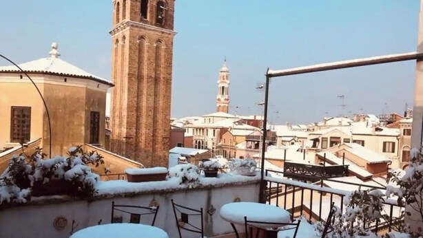 Una terraza en la azotea con mobiliario cubierto de nieve y vistas a una torre histórica y edificios circundantes.