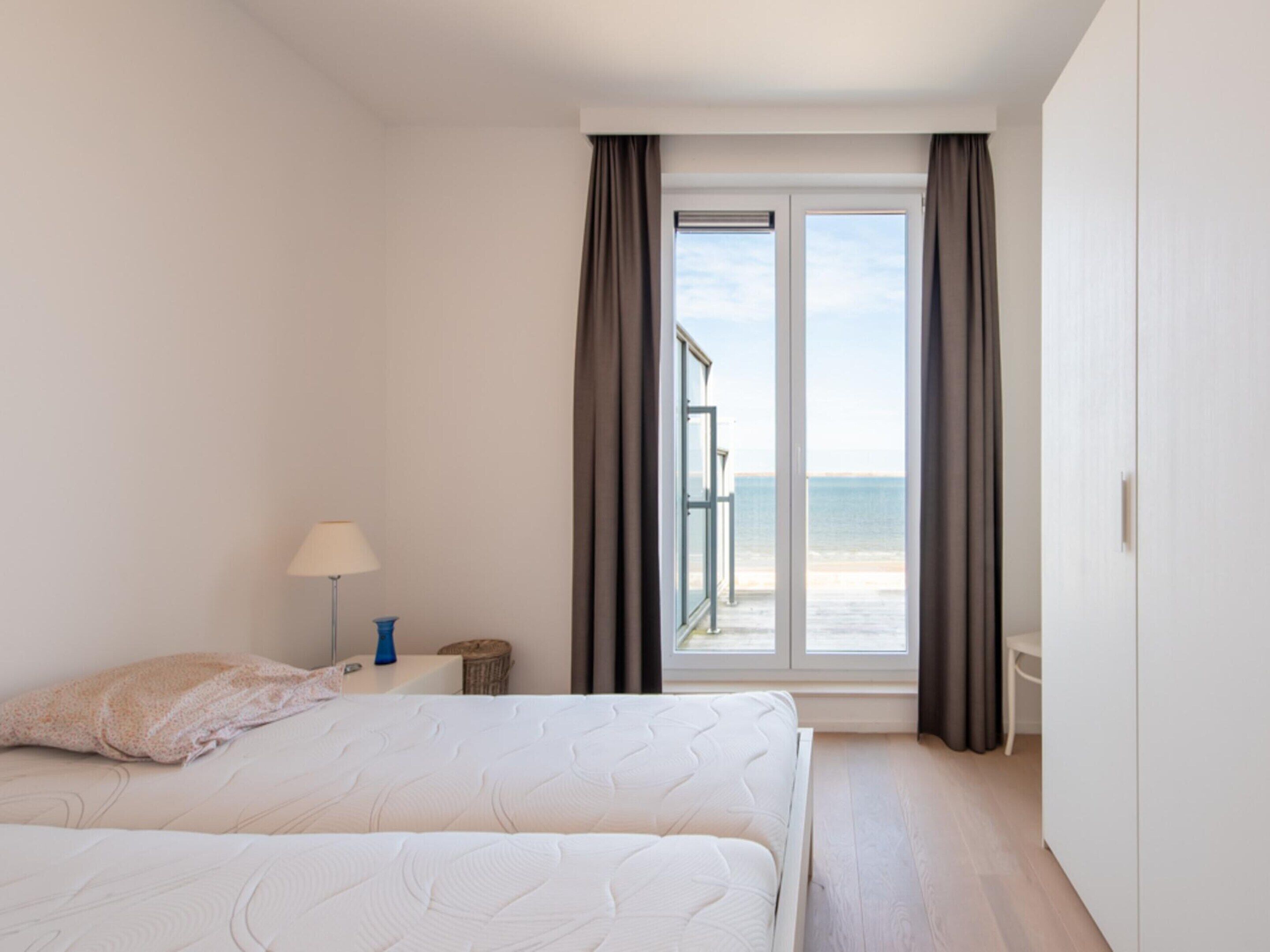 A bedroom with a bed, a nightstand, a lamp, and a view of the sea through a large window.