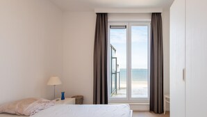 Un dormitorio con cama, mesita de noche, lámpara y vista al mar a través de un gran ventanal.