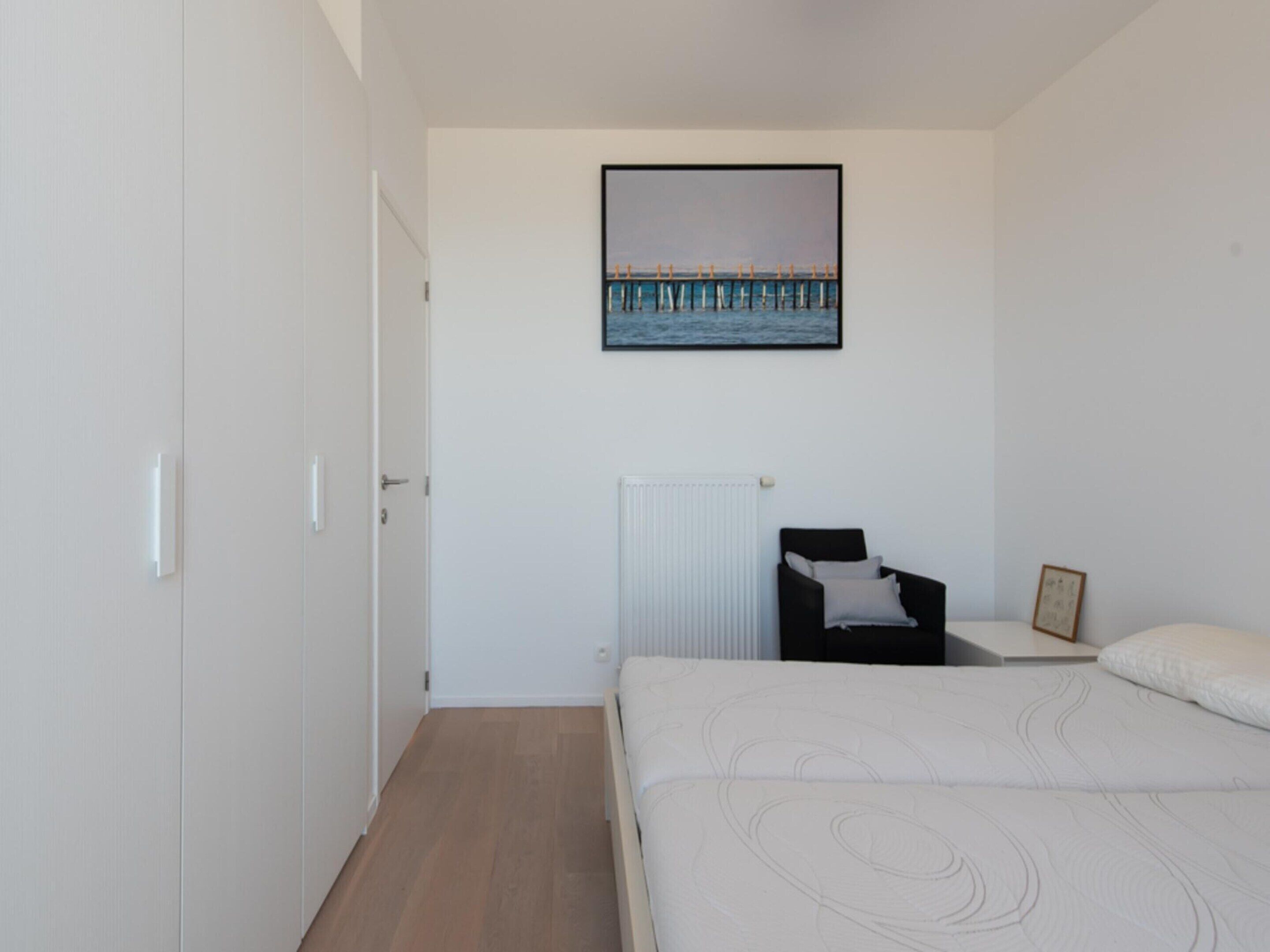 A bedroom with a bed, a chair, a radiator, and a picture of a pier on the wall.