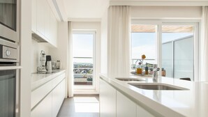 Una cocina moderna con armarios blancos, horno empotrado, cafetera y vistas a un paisaje urbano a través de un ventanal grande.