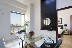 A modern hotel room with a glass dining table, a bed with a black headboard, a small bedside table with a lamp, and a view of a Ferris wheel through a large window.