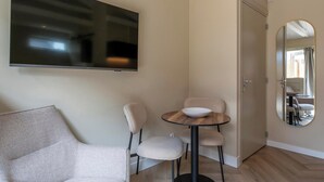 A modern living room with a flat-screen TV mounted on the wall, a round wooden table with a bowl, two upholstered chairs, and a large mirror on the wall. - Beautiful Suite on the Village Square (Callantsoog)