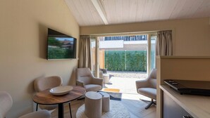 A modern living room with a flat-screen TV, comfortable seating, a dining table, and a view of a garden through large windows. - Beautiful Suite on the Village Square (Callantsoog)