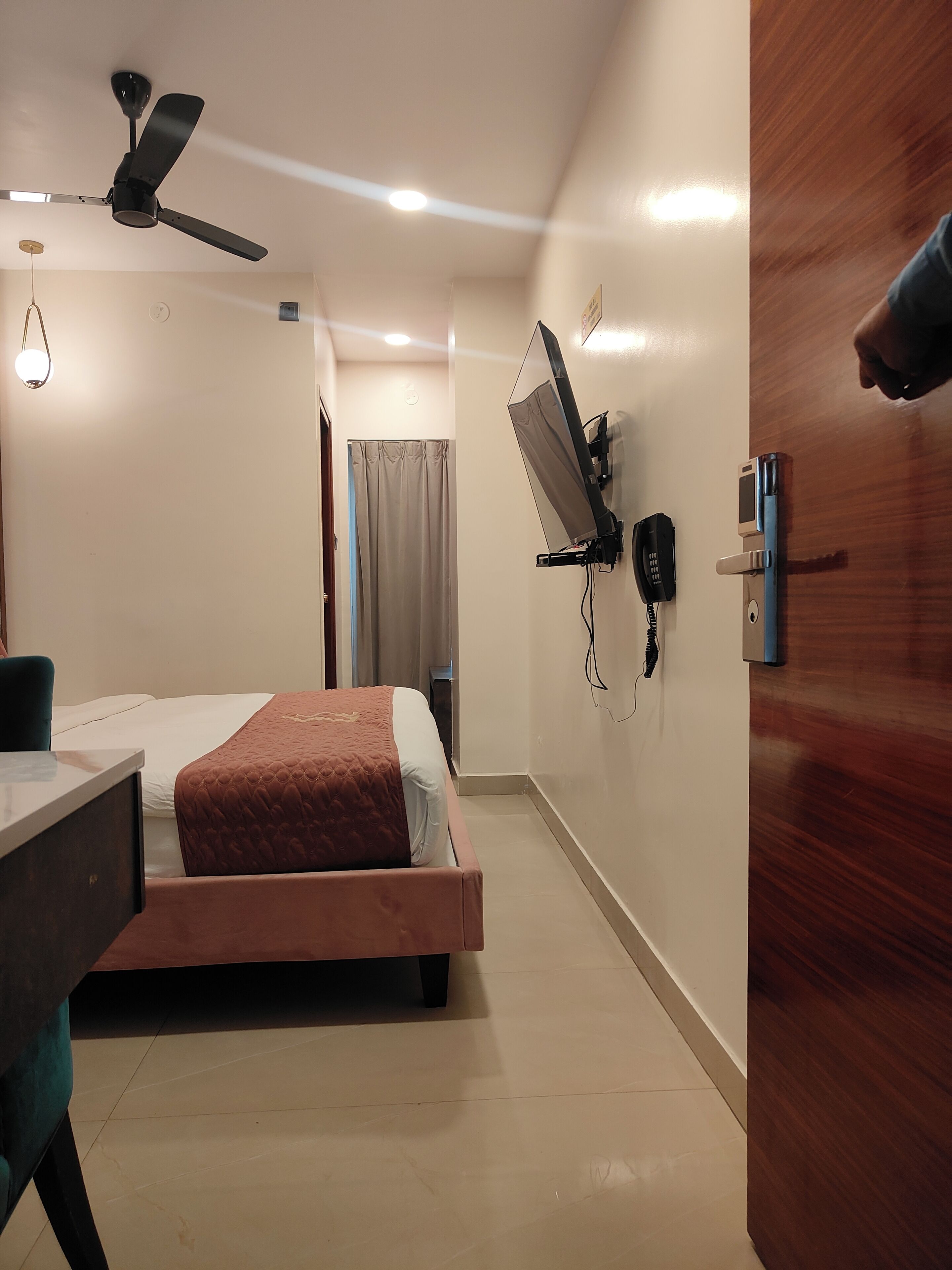 Ein Hotelzimmer mit Bett, Schreibtisch, Deckenventilator, Wandfernseher und einer Tür.