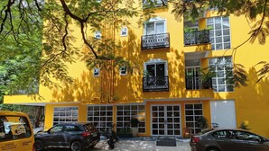Ein gelbes Gebäude mit zahlreichen Fenstern und Balkonen, ein Parkplatz mit Autos und ein Baum im Vordergrund.