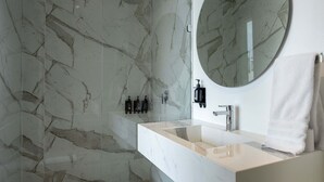 Ein modernes Badezimmer mit einer Duschwand aus Marmor, einem runden Spiegel, einem weißen Waschbecken und einem Handtuchhalter.