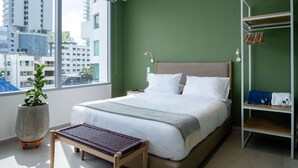 Ein modernes Hotelzimmer mit einem großen Bett, einem Nachttisch, einer Kommode, einer Zimmerpflanze, einer Bank, einem Kleiderschrank und Blick auf die Stadt.