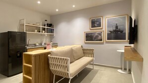 A compact living space with a kitchenette, a sofa, a wooden side table, and a flat-screen TV mounted on the wall.