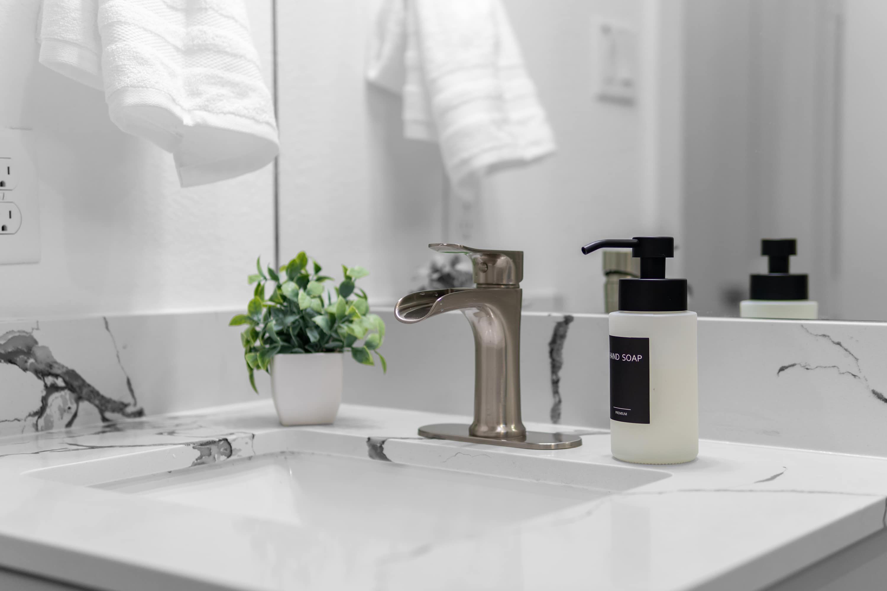 Un baño con lavamanos de mármol blanco, un grifo moderno, una planta en maceta y una botella de jabón para manos.