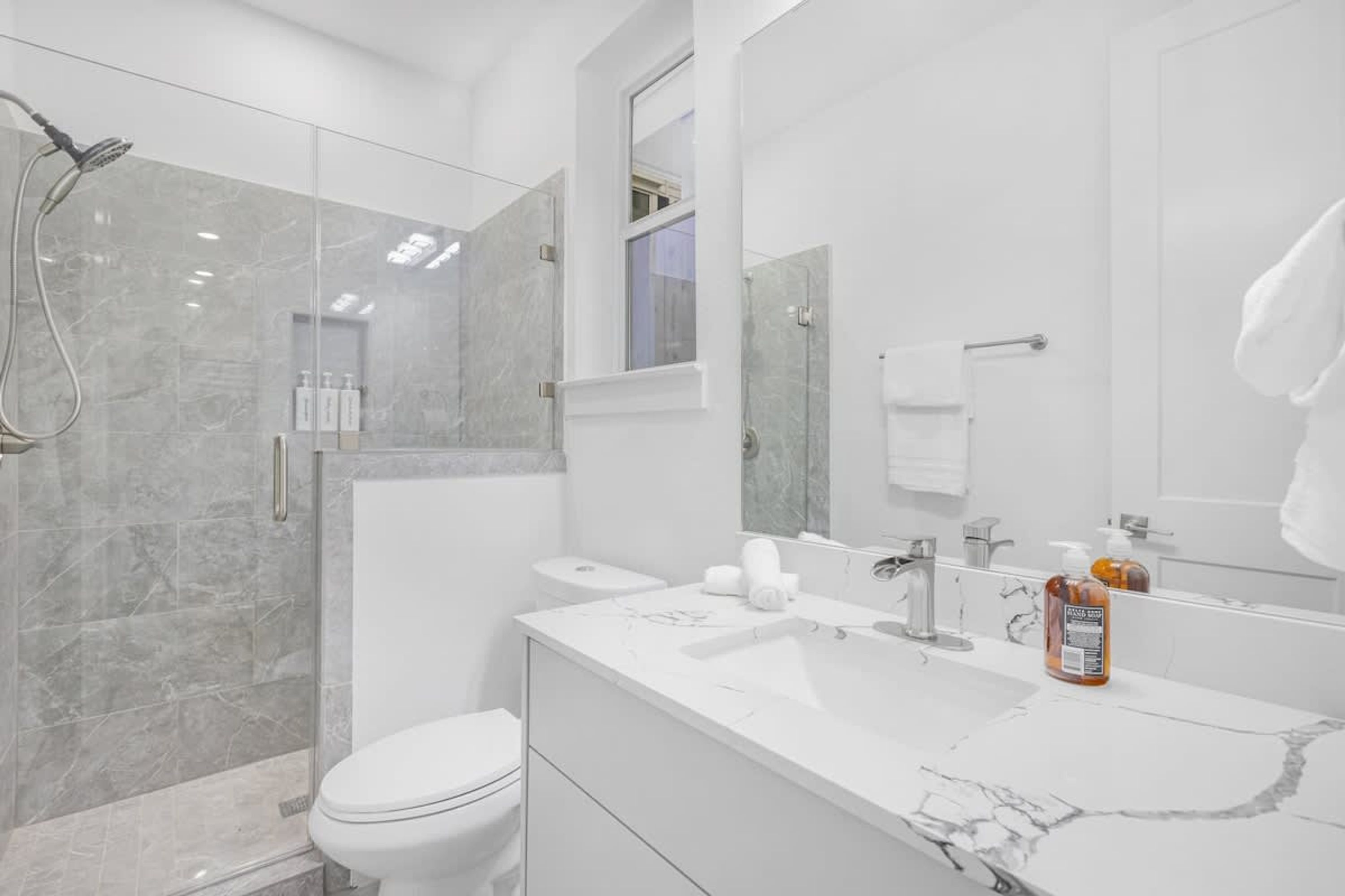 A modern bathroom with a glass-enclosed shower, a white toilet, a marble sink, and a large mirror.