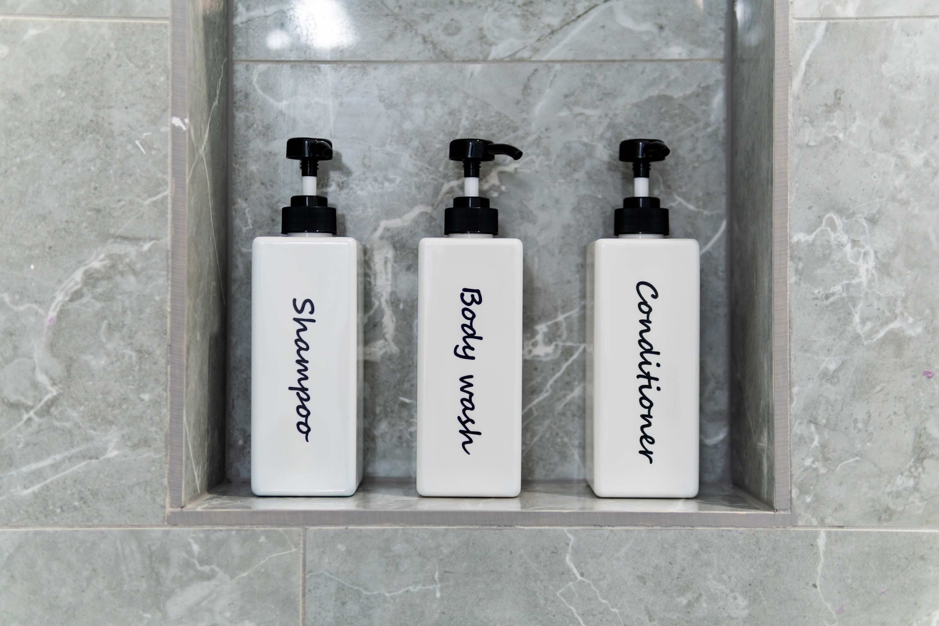 Tres botellas blancas con dosificadores negros etiquetadas 'Champú', 'Gel de ducha' y 'Acondicionador' en un baño.
