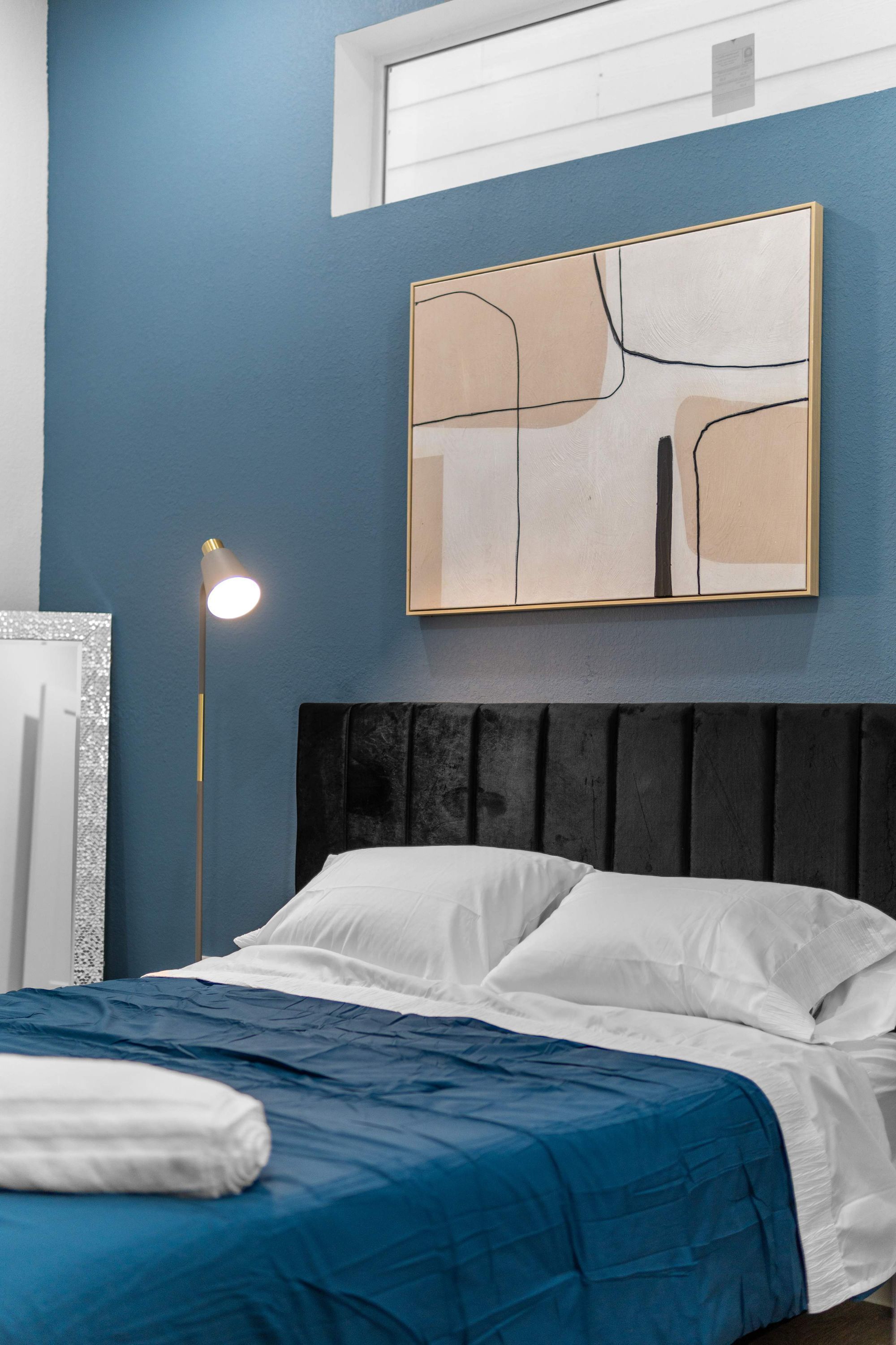 A neatly made bed with a blue and white bedspread, a black headboard, and a framed abstract painting on the wall.