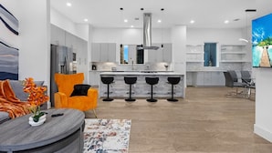 Una cocina moderna con una isla central, taburetes de bar y un comedor con una mesa redonda y una silla naranja.