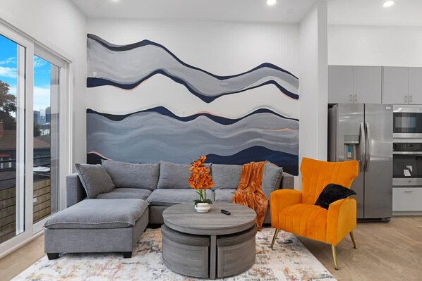A modern living room with a grey sectional sofa, an orange armchair, a grey coffee table, and a large abstract wall mural. - Spacious Houston 3bed3 Bath Family Townhome (Houston)