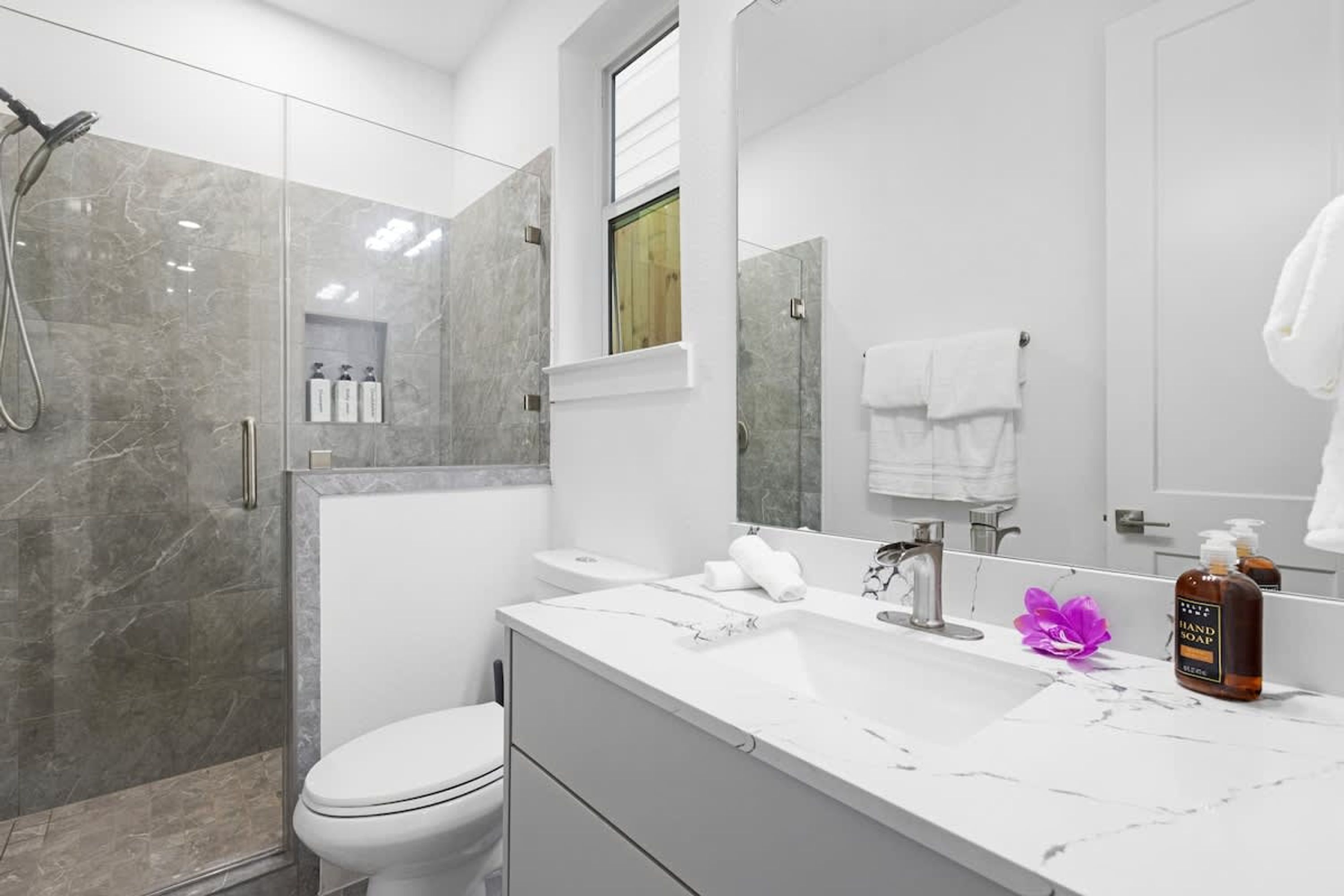 Un baño moderno con ducha de vidrio, inodoro blanco, lavabo de mármol y un espejo grande.