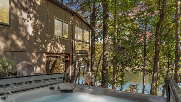 A hot tub with a view of a lake and trees, a wooden deck, and a building with large windows.