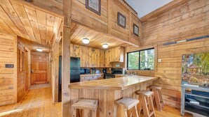 A wooden cabin interior with a central kitchen, bar area, and a large window overlooking a forest.