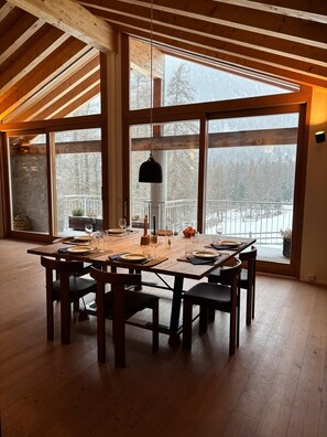 Ein Essbereich mit einem großen Holztisch, gedeckt für vier Personen, großen Fenstern mit Blick auf eine verschneite Landschaft und einer hängenden Pendelleuchte.