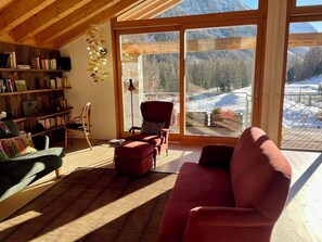 Ein gemütliches Wohnzimmer mit einem roten Sofa, einem Holzregal und Blick auf eine verschneite Landschaft durch große Glasfenster.