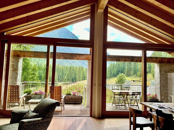 Ein geräumiges Zimmer mit großen Fenstern und Blick auf die Berge, Holzbalken an der Decke sowie ein Balkon mit Sitzgelegenheit im Freien.
