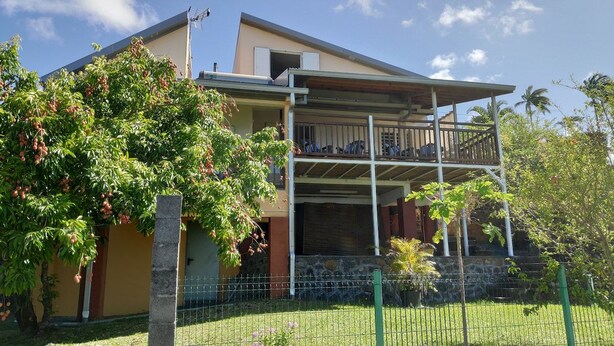 Una casa a due piani con un balcone, circondata dal verde e un cortile recintato.