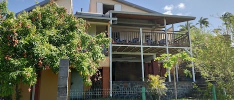 Una casa de dos pisos con balcón, rodeada de vegetación y un patio cercado.