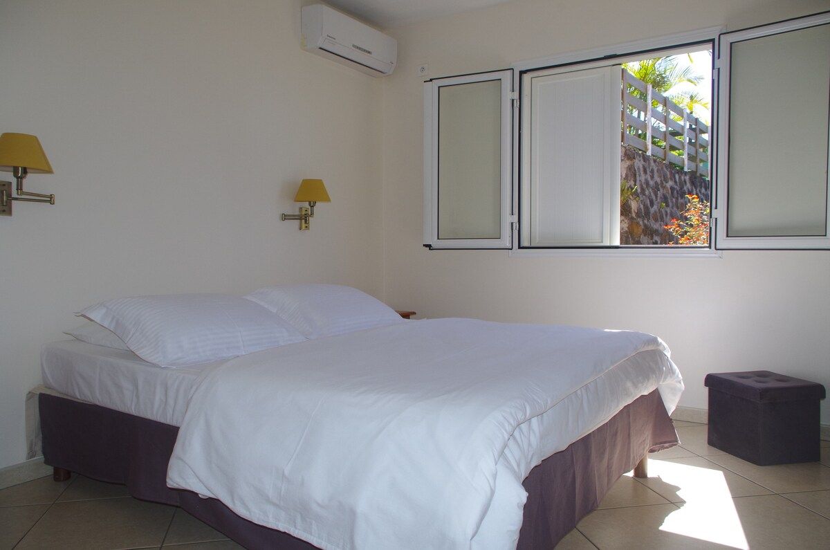 A bedroom with a bed, two wall-mounted lamps, an air conditioning unit, and a window with a view of a white fence and greenery.