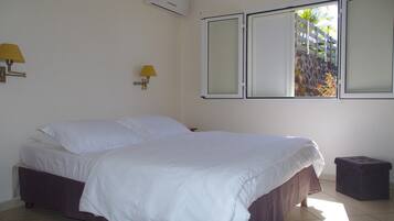 A bedroom with a bed, two wall-mounted lamps, an air conditioning unit, and a window with a view of a white fence and greenery.