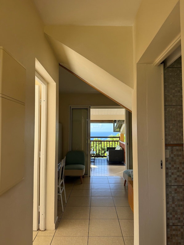 A corridor leading to a balcony with a view of the ocean, a chair, and a sofa.