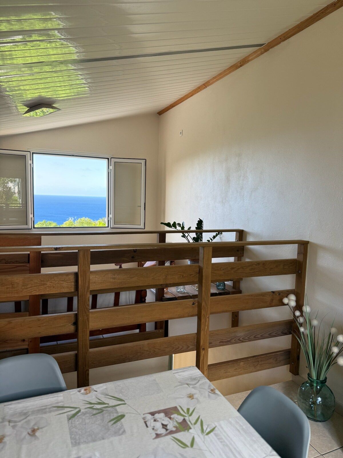 A room with a view of the ocean, a wooden partition, a table with a floral tablecloth, and a vase with white flowers.