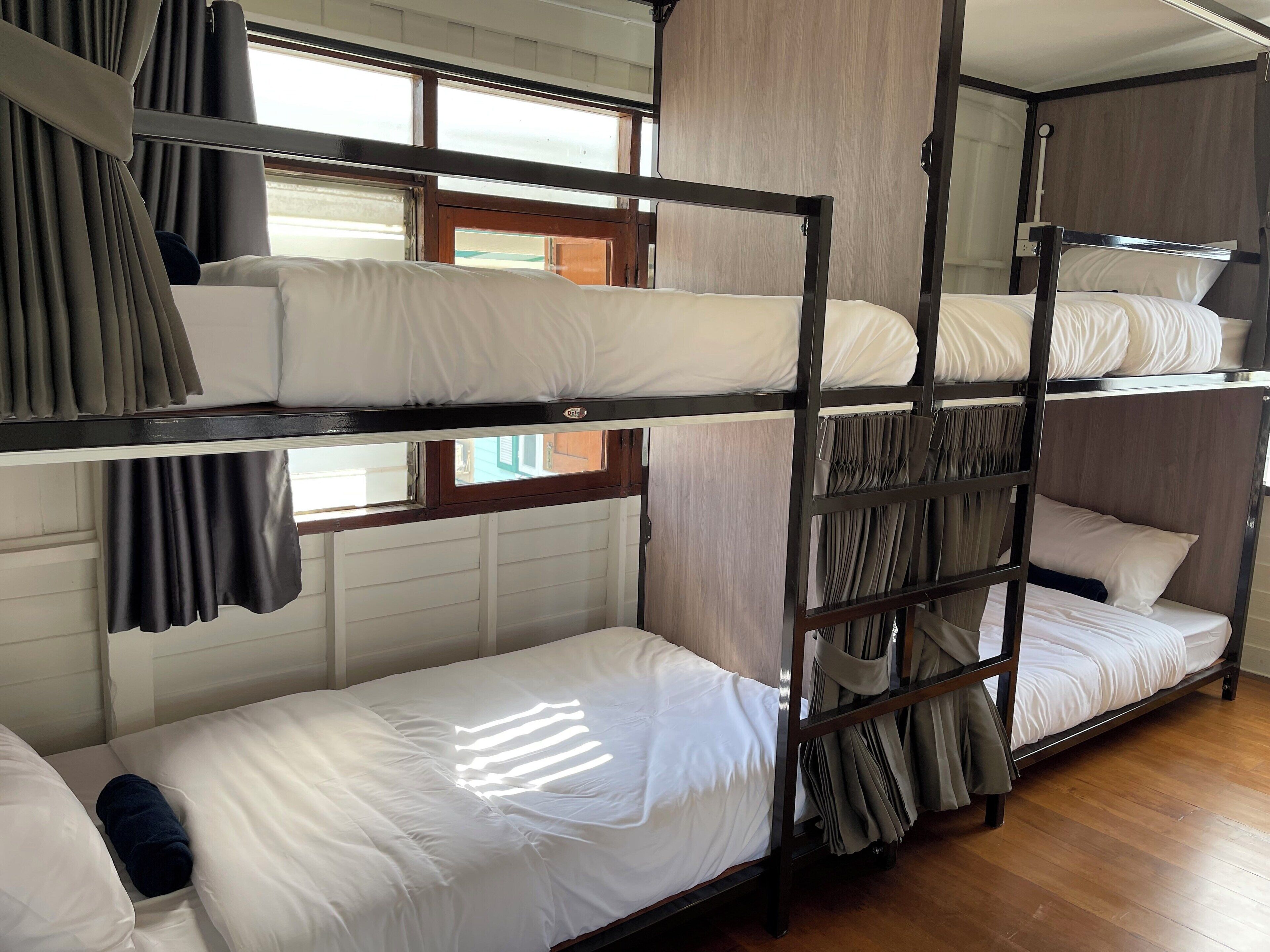 Une chambre avec un lit superposé, du linge de lit et des rideaux blancs, un plancher en bois et une fenêtre également dotée de rideaux.