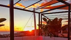Ein moderner Außenbereich mit einer weißen Couch und einem kleinen Tisch, mit Blick auf einen Sonnenuntergang über dem Meer.
