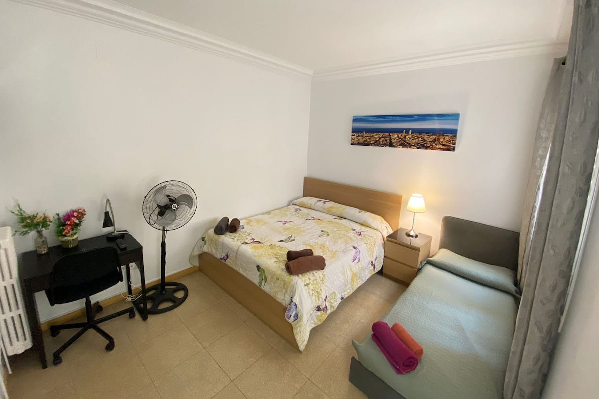 A hotel room with a bed, a desk, a chair, a fan, a nightstand with a lamp, and a painting on the wall.