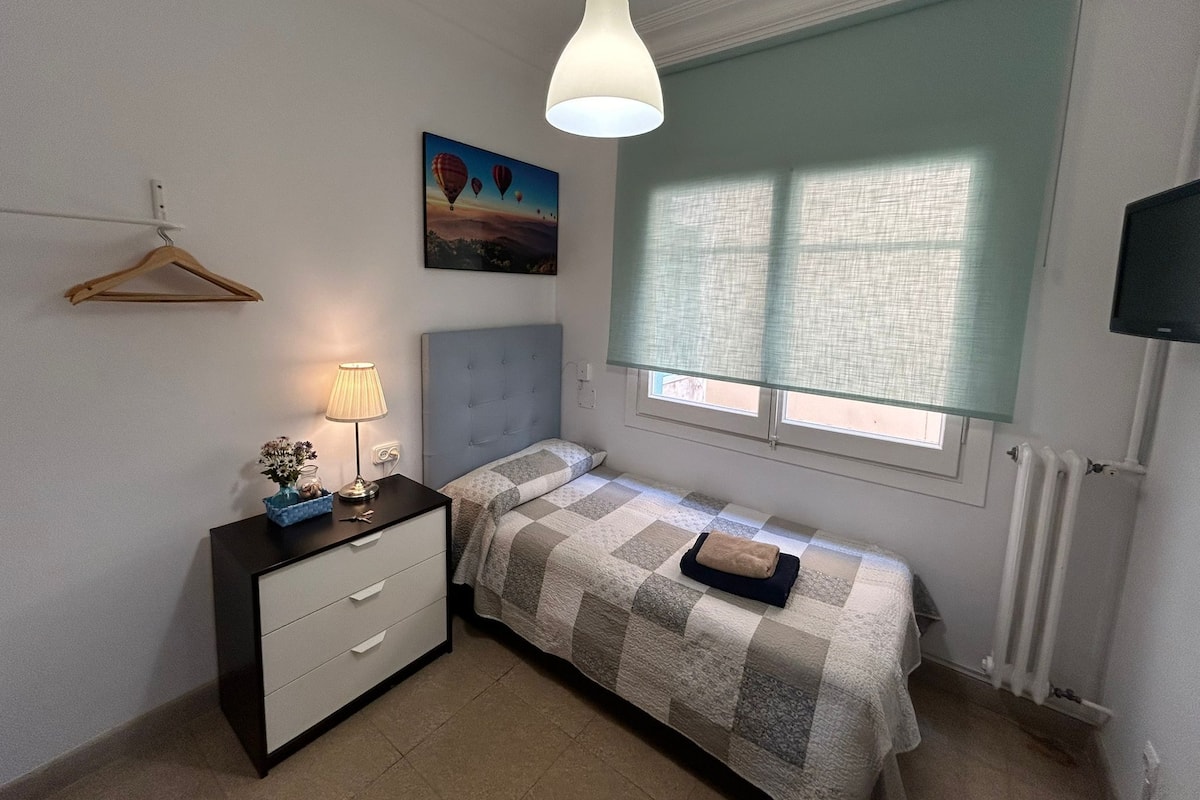 A small bedroom with a single bed, a bedside table with a lamp, a hanging light fixture, a window with blinds, and a painting on the wall.