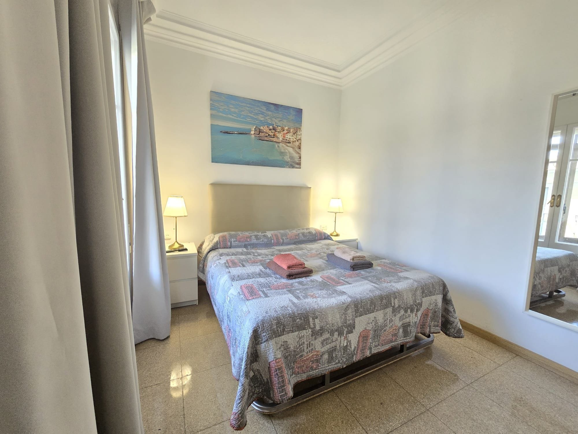 A hotel room with a bed, two bedside lamps, a painting of a coastal scene, and a large mirror.
