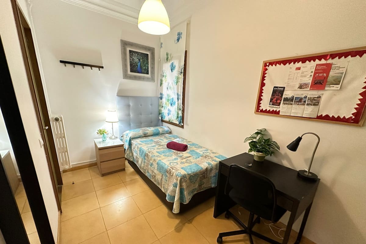 A small, tidy bedroom with a bed, a desk, a chair, a radiator, a wall-mounted shelf, and a bulletin board with papers.