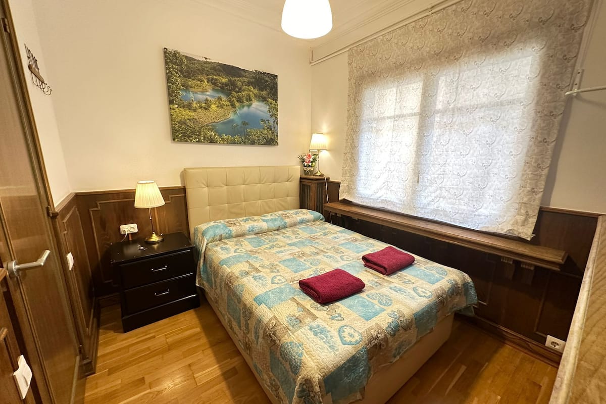 A bedroom with a bed, bedside tables, a lamp, a painting, and a window with curtains.