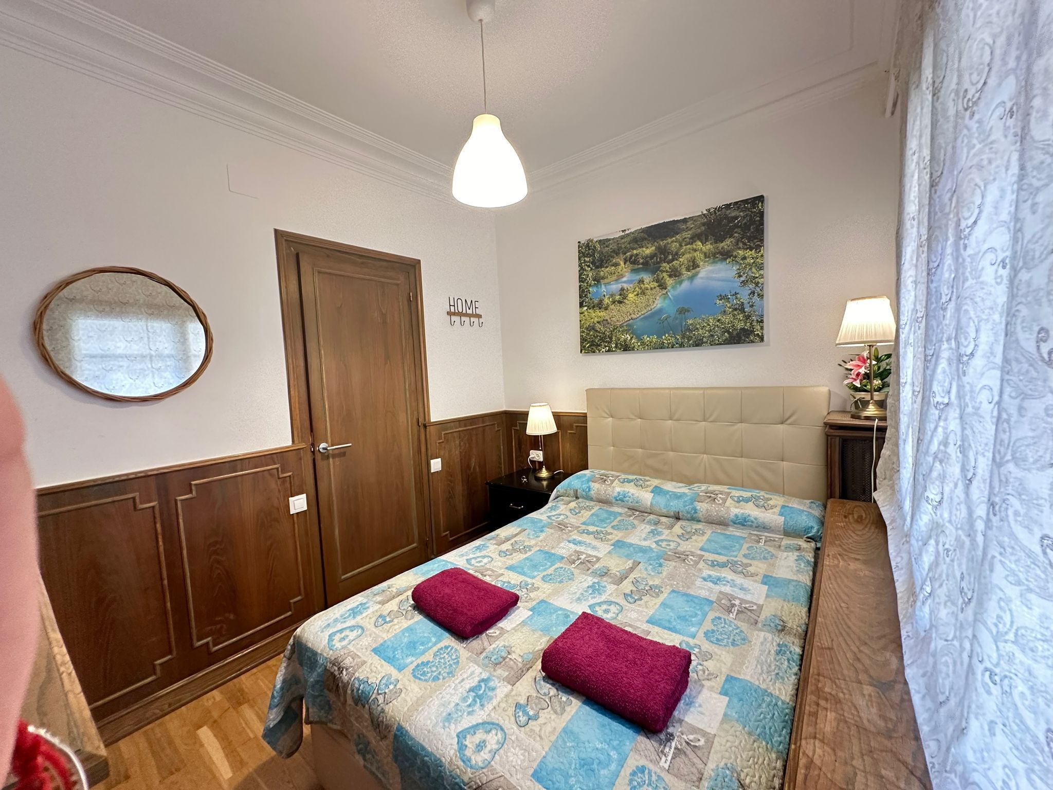 A bedroom with a bed, a mirror, a door, a painting, and a nightstand with a lamp.