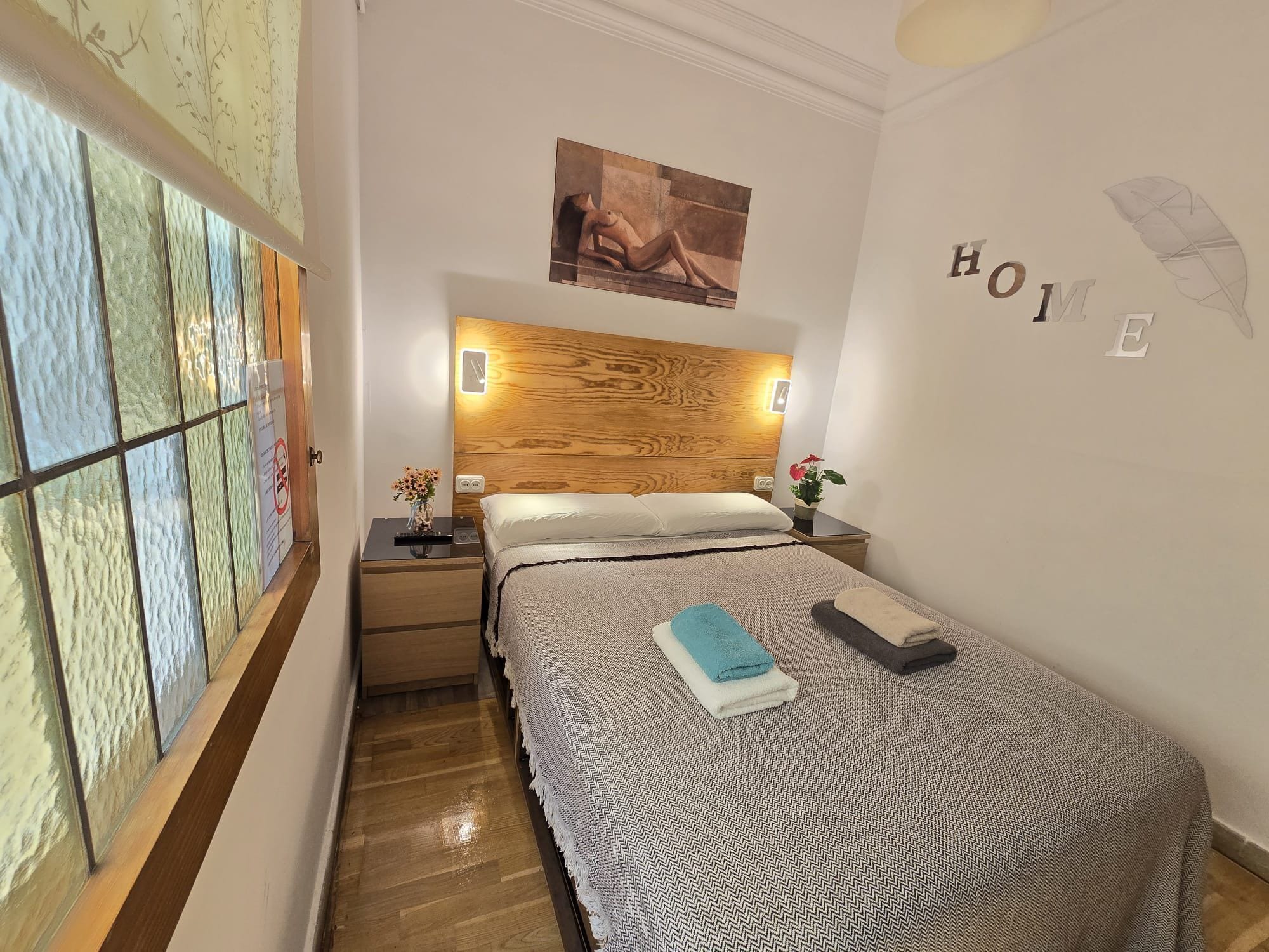 Une chambre à coucher bien aménagée, avec une tête de lit en bois, un lit recouvert d’une couvre-lit à motifs, une table de chevet ornée d’un vase de fleurs et une œuvre d’art accrochée au mur.