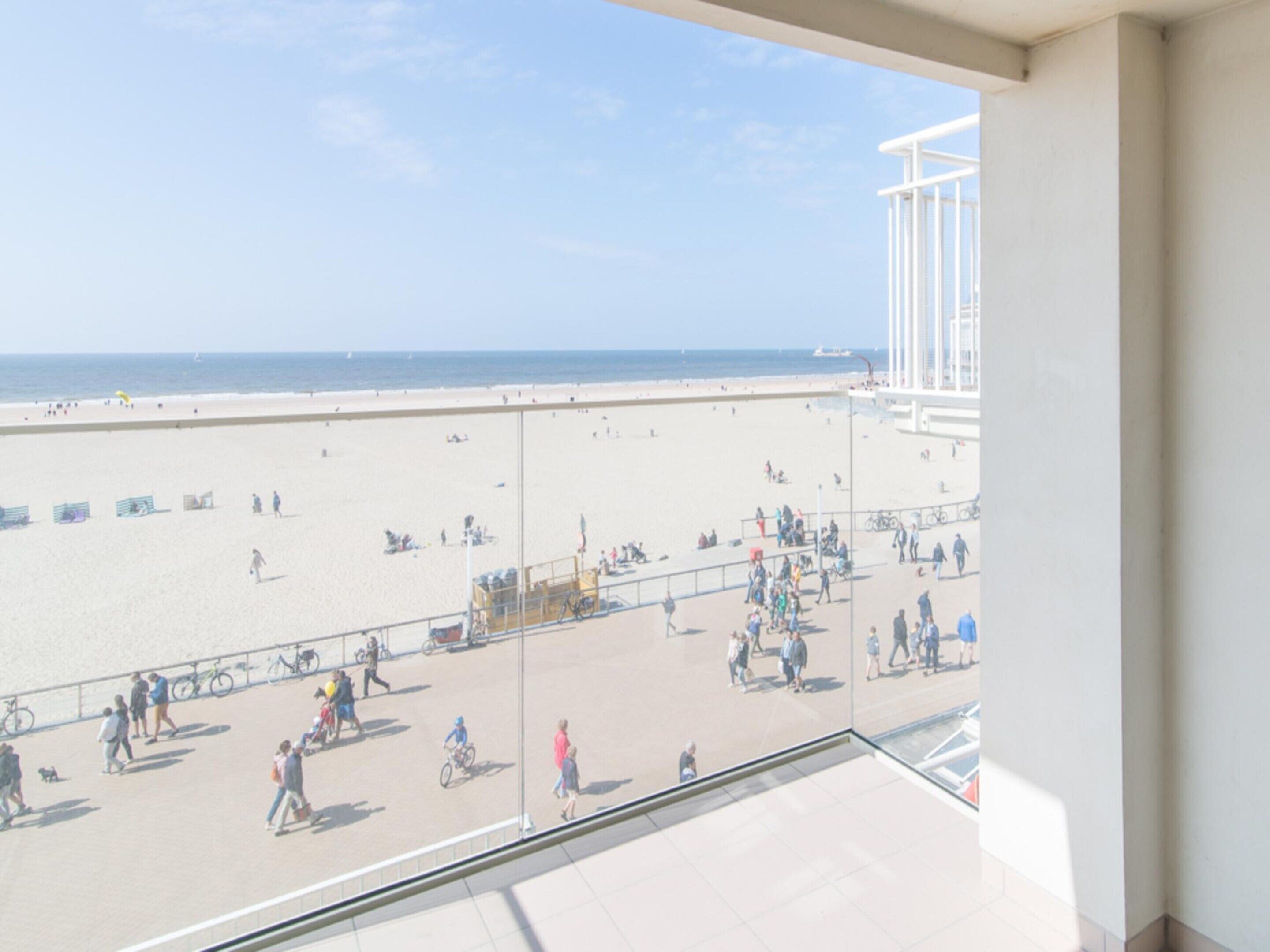 Blick auf einen Strand vom Balkon aus, mit blauem Himmel, Sandstrand und Menschen, die spazieren gehen und Fahrrad fahren.