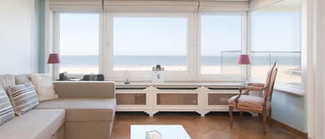 A spacious living room with a large window overlooking the beach, a sofa, a chair, and a coffee table with decorative items.