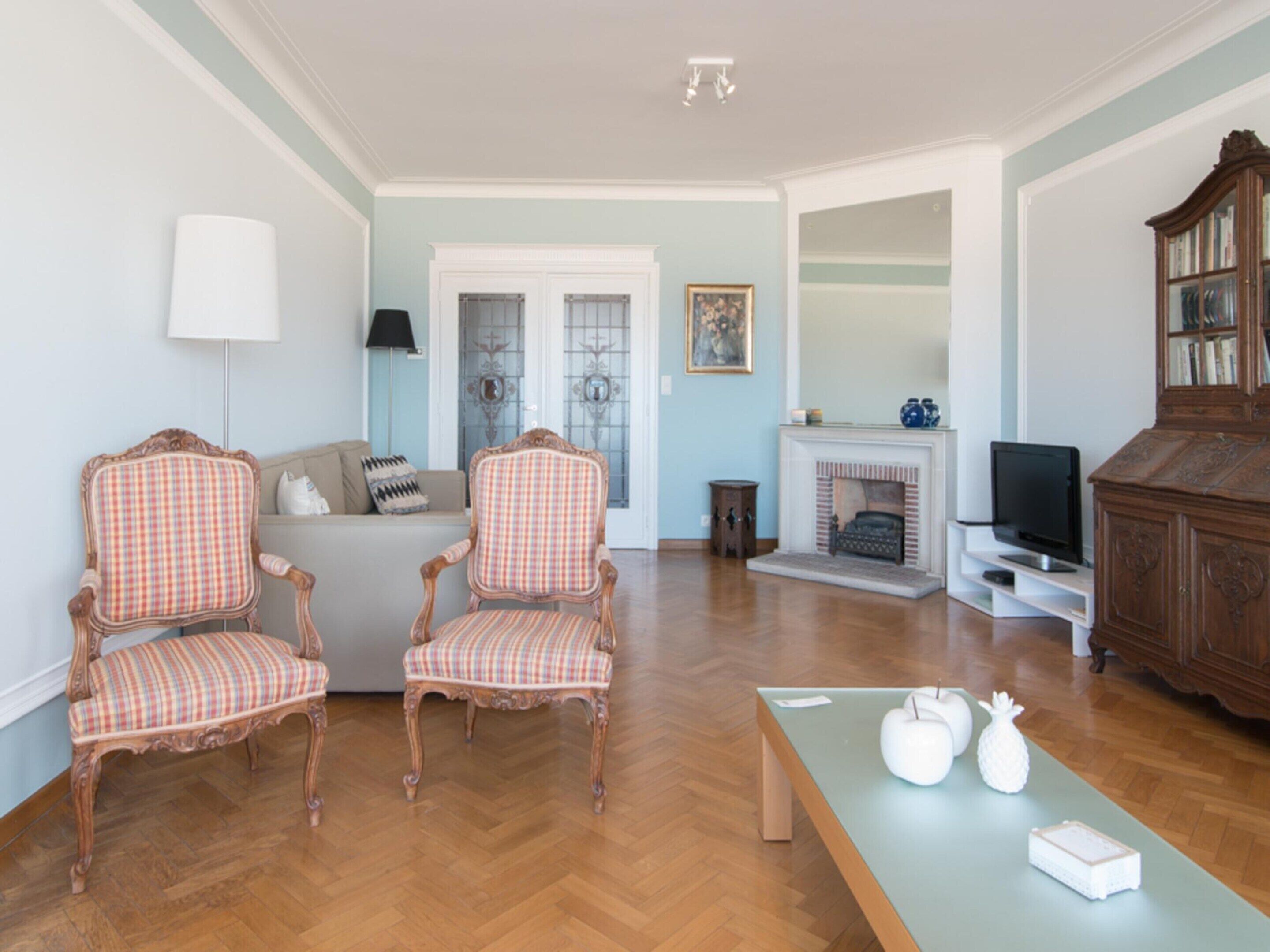 A living room with two ornate chairs, a sofa, a fireplace, a television, and a bookshelf.