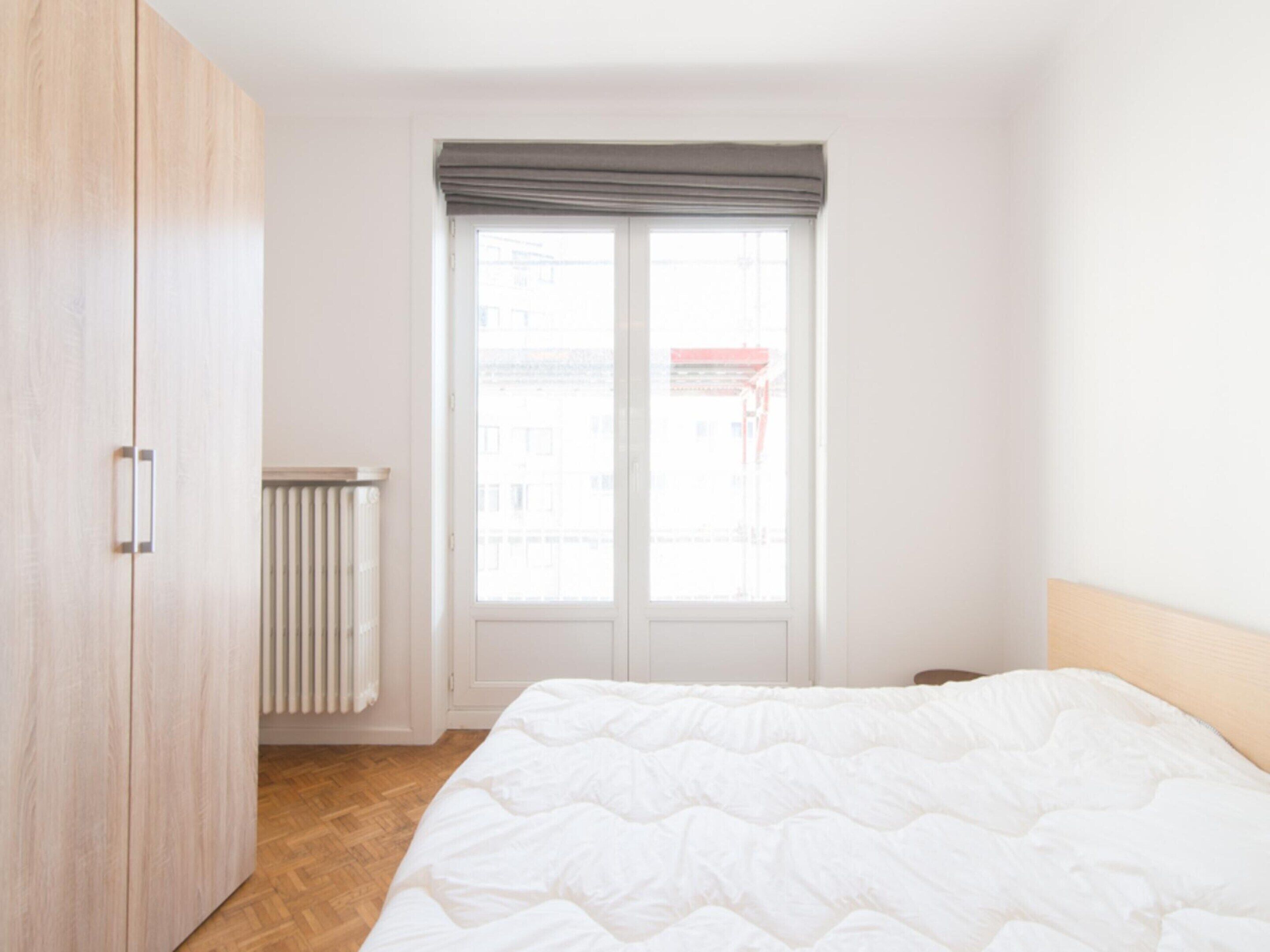 A bedroom with a bed, a wardrobe, a radiator, and a window with a view of buildings.